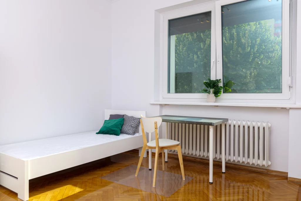 Minimalistyczny pokój z łóżkiem i biurkiem przy oknie, parkiet i zieleń widoczna za szybą