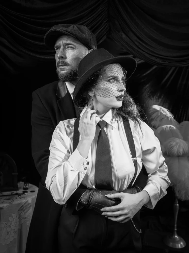 Czarno-biały portret pary w stylizacji noir - kobieta w kapeluszu z woalką przed mężczyzną w kaszkiecie, zasłony w tle
