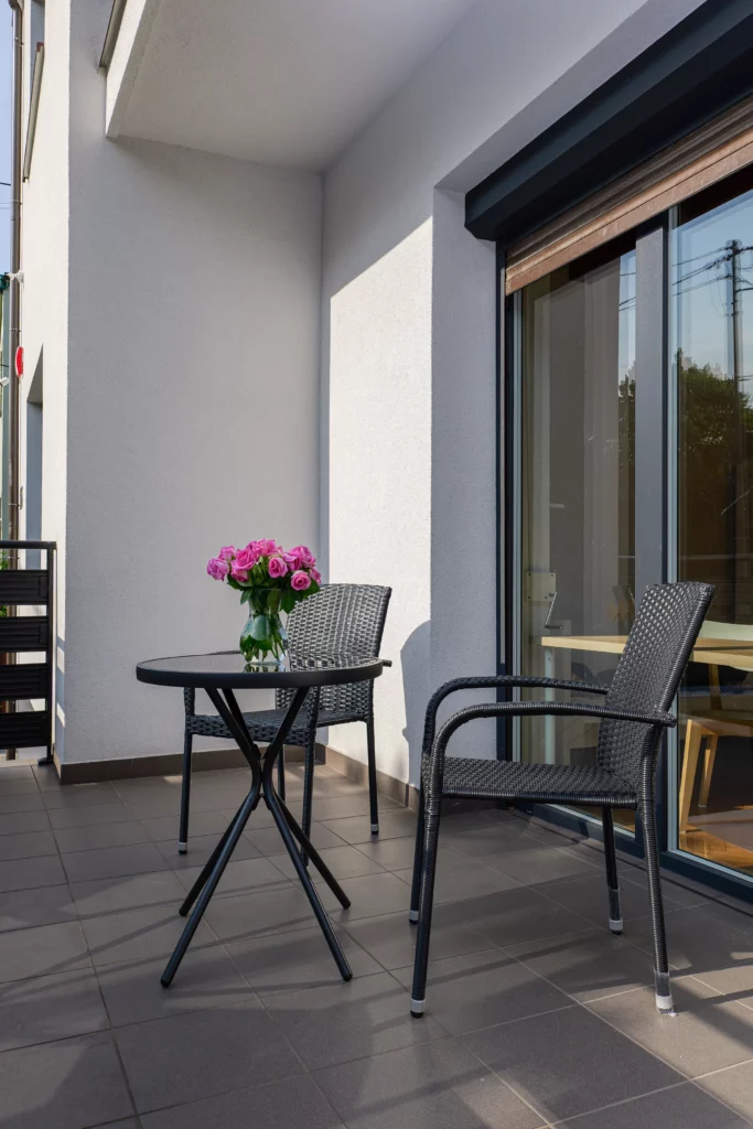 Niewielki balkon z okrągłym stolikiem i wiklinowym fotelem, róże na blacie, boczne światło