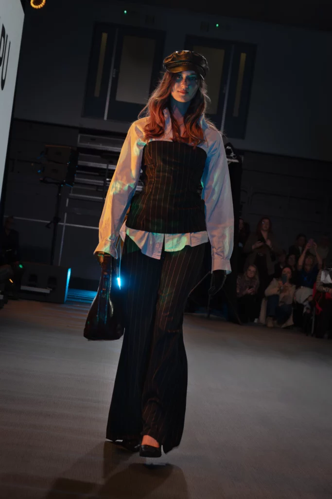 Modelka w prążkowanym gorsecie na białej koszuli i szerokich spodniach, skórzana czapka, czarna torba, androgyniczny look