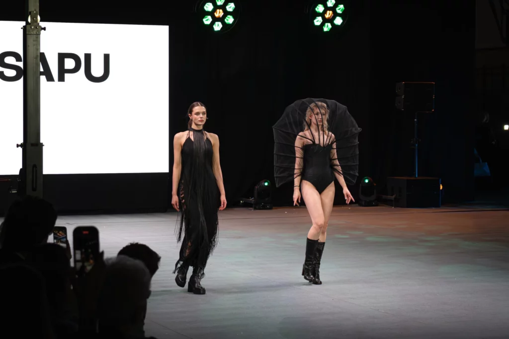 Dwie modelki w czerwono-czarnych satynowych sukniach z gorsetami i koronkowymi maskami na twarzach, długie treny na scenie
