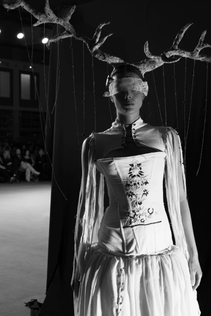 Modelka z porożem na głowie i koronkową opaską na oczach, biały haftowany gorset, czarno-biała sceneria z koralikami