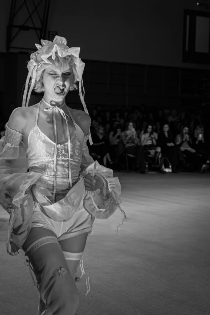 Modelka w białej falbaniastej stylizacji z kokardami i wstążkami, ekspresyjna mina, czarno-biała scena, publiczność w tle